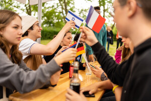 Mehrere Jugendliche sitzen an einem Biertisch unter einem Zelt. Sie halten kleine Deutschland-, Fankreich- und Israelfahnen. Die Jugendlichen lächeln und trinken Softdrinks.