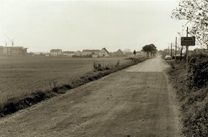 Ein Schotterweg, links ein großes Feld. Auf der rechten Seite des Weges steht das Ortsschild von Monheim
