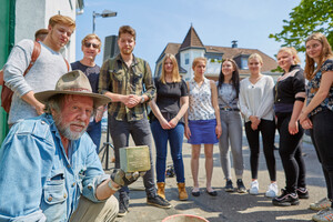 Gunter Demnig hält einen Stolperstein hoch, im Hintergrund steht eine Gruppe Schülerinnen und Schüler