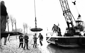 Ein Schiff mit einem Kran am Rheinufer. Am Kran hängt eine Fünf-Zentner-Bombe