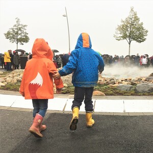 Zwei Kinder in Regenkleidung vor dem Monheimer Geysir mit dem Rücken zur Kamera.