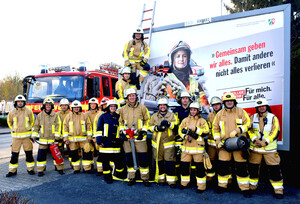 Eine Gruppe von Feuermännern und -frauen vor einem Einsatzwagen und einem Plakat mit Webrung für die Feuerwehr