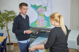 Ein junger Mann zeigt einer Frau etwas auf einem Tablet, im Hintergrund ist eine Karte der Stadt Monheim am Rhein