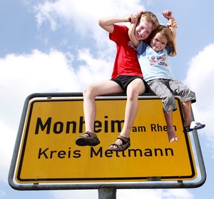 Gelbes Ortsschild von Monheim am Rhein vor blauem Himmel. Auf dem Schild sitzen zwei Kinder.