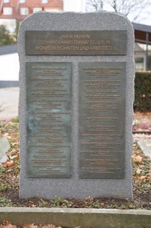 Ein Stein mit mehreren Metallplatten, auf der oberen Platte steht: "Hier ruhen Zwangsarbeitskräfte, die in Monheim wohnten und arbeiteten"