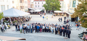 Gruppenfoto von Vertreterinnen und Vertretern Monheim am Rhein vor dem Rathaus in Wiener Neustadt. Im Hintergrund sitzen Menschen unter Sonnenschirmen vor Restaurants.  