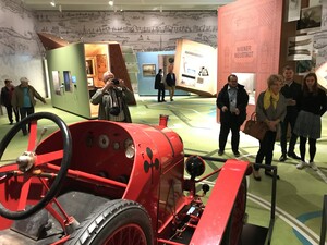 Mehrere Besucher in einem Ausstellungsraum, links im Vordergrund steht ein roter Oldtimer