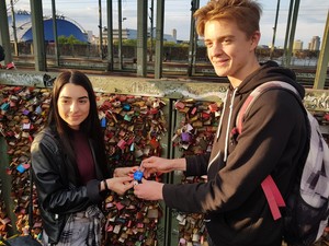 Ein Schüler und eine Schülerin halten gemeinsam ein kleines Schloss vor den vielen Liebesschlössern an der Kölner Hohenzollernbrücke