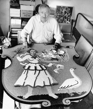 Hans Schweizer an einem Tisch. Vor ihm das große Mosaik mit Gänseliesel und Gans, in das er Mosaiksteine einfasst