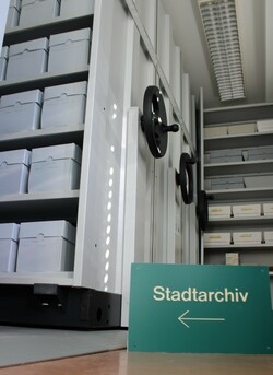 Blick in das Stadtarchiv: In der Mitte ein grünes Schild mit der Aufschrift Stadtarchiv, links daneben mehrere Regale mit Kisten