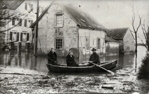 Altes schwarz-weiß Foto: drei Menschen auf einem Ruderboot, im Hintergrund viele vom Wasser angegriffene Häuser mit erheblichen Schäden