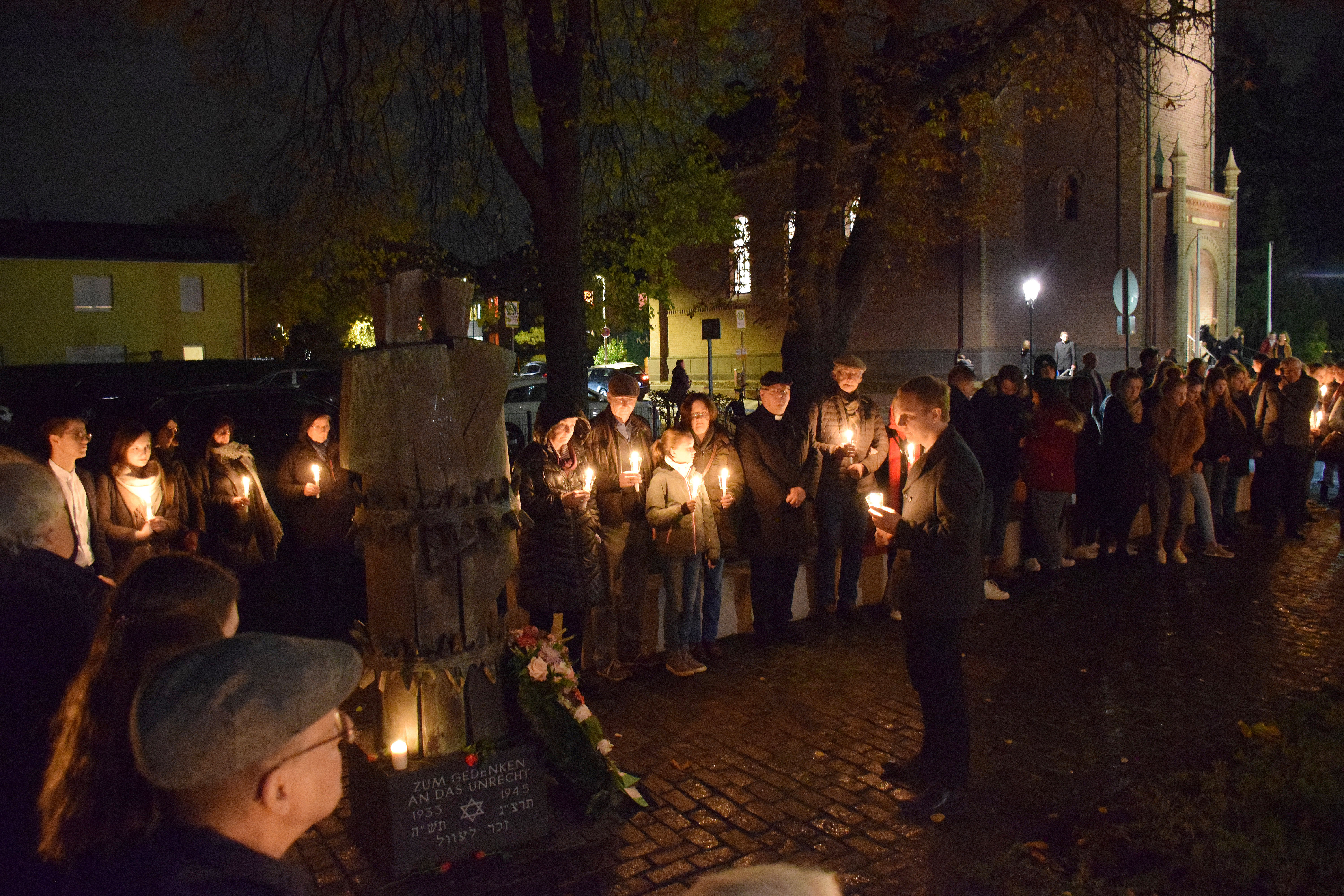 Eine Gruppe Menschen am Mahnmal am Kradepohl. Alle halten Kerzen, es ist dunkel.