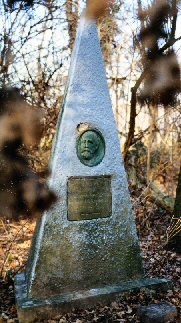 Ein dreieckiger Gedenkstein mit einer ovalen Platte und dem herausgearbeiteten Gesicht von Clemens Kleinn und einer goldenen Tafel