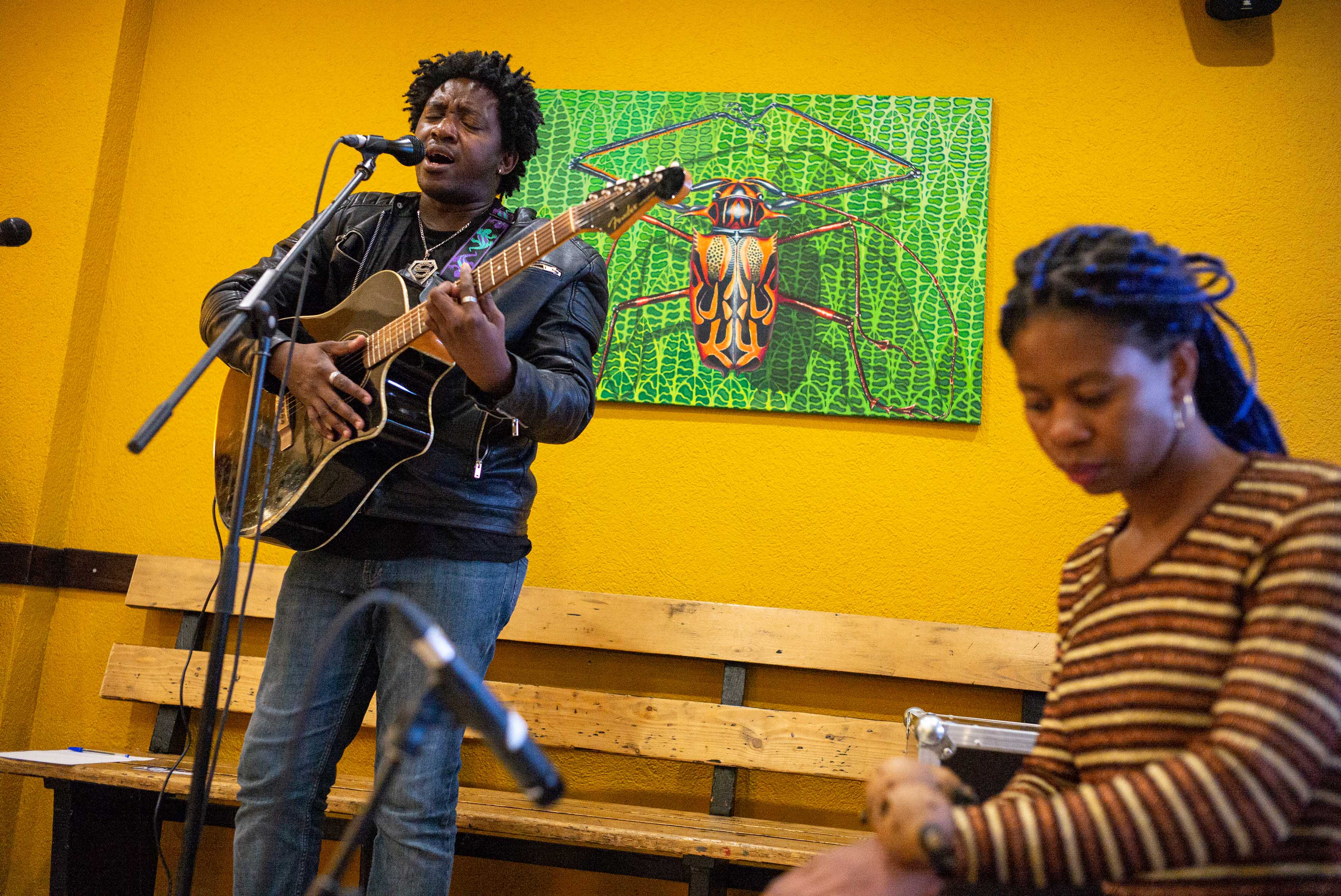 Ein Mann mit einer Gitarre singt in ein Mikrofon, neben ihm sitzt eine Frau